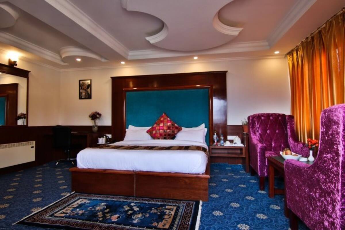 Una habitación de hotel con una cama grande, un cabecero de madera, un sillón morado, una mesita pequeña y una alfombra azul estampada.