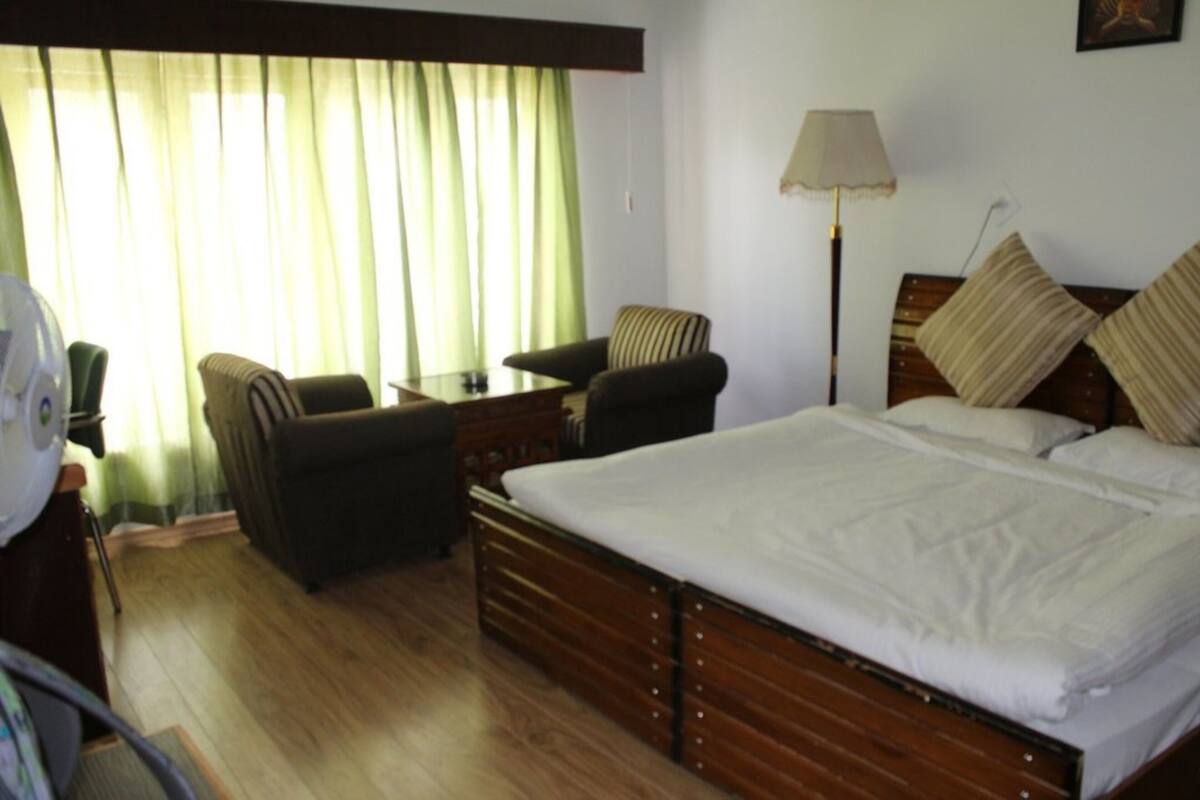Habitación de hotel con una cama grande, dos sillones, una mesita, una lámpara y una ventana con cortinas.