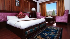 A hotel room with a large bed, a desk, two chairs, a window with a mountain view, and a patterned rug. - TIH Hotel Shangrila-Ladakh (Leh)
