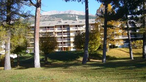 A multi-story hotel with a flat roof and balconies, surrounded by trees and a grassy lawn. - Charming T2 in Barcelonnette with terrace for 4 people (Barcelonnette)