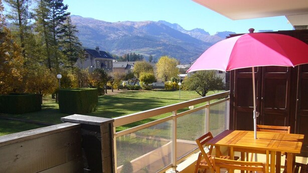 A balcony with a wooden table and chairs, a red umbrella, and a view of a green lawn and mountains. - Charming T2 in Barcelonnette with terrace for 4 people (Barcelonnette)
