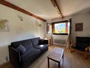 A living room with a sofa, a wooden coffee table, a chair, a window with a view, a lamp, and a radiator. - Spacious T2 facing slopes - 6 pers, South, Comfort (Enchastrayes)