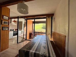 A room with a bed, a wooden floor, a kitchen area, and a view of a green landscape through a glass door. - Spacious T2 facing slopes - 6 pers, South, Comfort (Enchastrayes)
