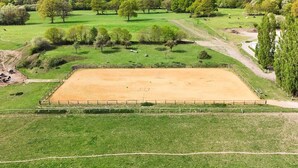A fenced horse riding arena surrounded by green fields and trees.