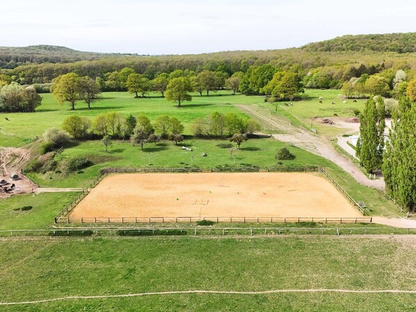 A fenced horse riding arena surrounded by green fields and trees.
