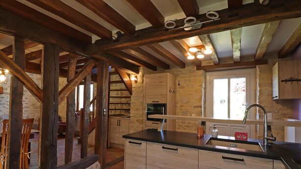 A rustic kitchen with exposed wooden beams, a stone wall, and a large window.