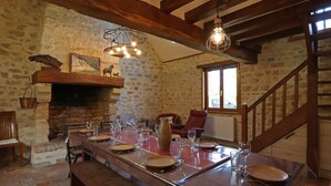 A rustic dining area with a stone wall, wooden beams, a fireplace, and a long table set for dining.