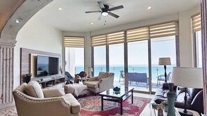 A spacious living room with a large TV, comfortable seating, a coffee table, and a view of the ocean.