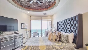 A bedroom with a large bed, a TV, a dresser, and a view of the ocean.