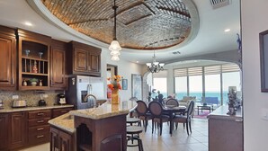 A spacious kitchen with dark wooden cabinets, a granite countertop, and a central island. There is a dining area with a large table and chairs, and a view of the ocean through large windows.