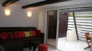 A living room with a dark-colored sofa, a small table, and a red ottoman. There are two wall-mounted lights and a sliding glass door leading to an outdoor area with a bamboo fence. - Charming Villa with Terrace, A/C, near Tantra Club, Private Parking (Cap d'Agde naturisme)