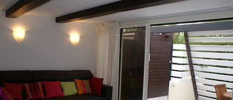 A living room with a dark-colored sofa, a small table, and a red ottoman. There are two wall-mounted lights and a sliding glass door leading to an outdoor area with a bamboo fence.