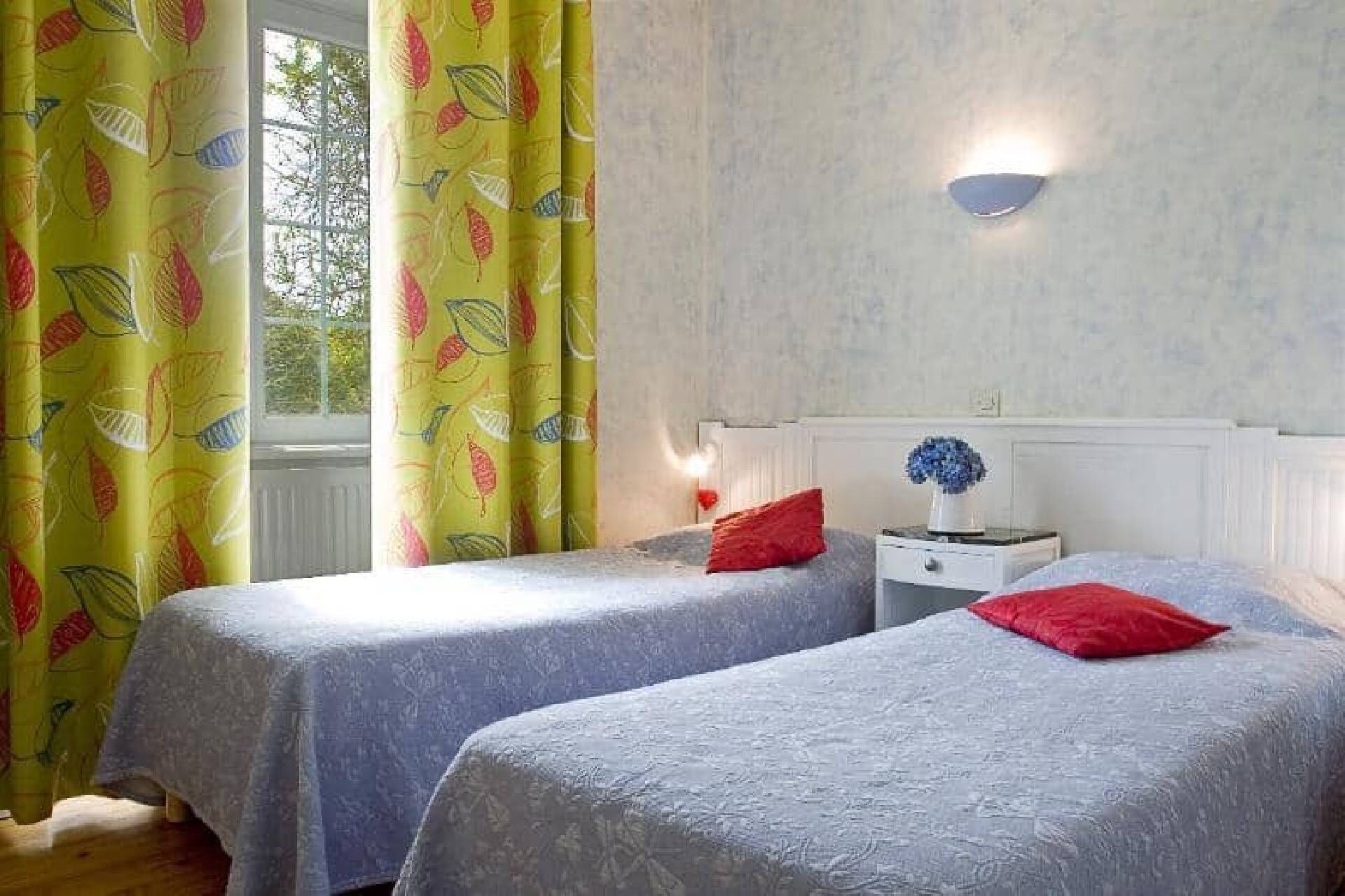 A room with two beds, a nightstand with a vase, and a window with curtains.