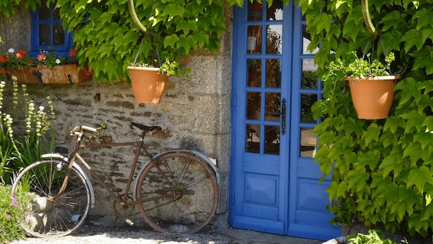 Una pared de piedra rústica con una puerta azul, una bicicleta y macetas colgantes con follaje.