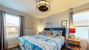Une chambre avec un grand lit, une table de chevet, un ventilateur fixé au mur et une fenêtre avec des rideaux.