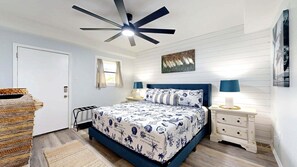 A bedroom with a bed, a ceiling fan, a window, a painting, and a nightstand.