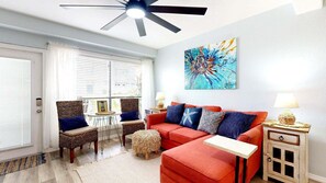 Un salon avec un canapé rouge, deux fauteuils, un ventilateur de plafond, un tableau et une petite table avec une lampe.