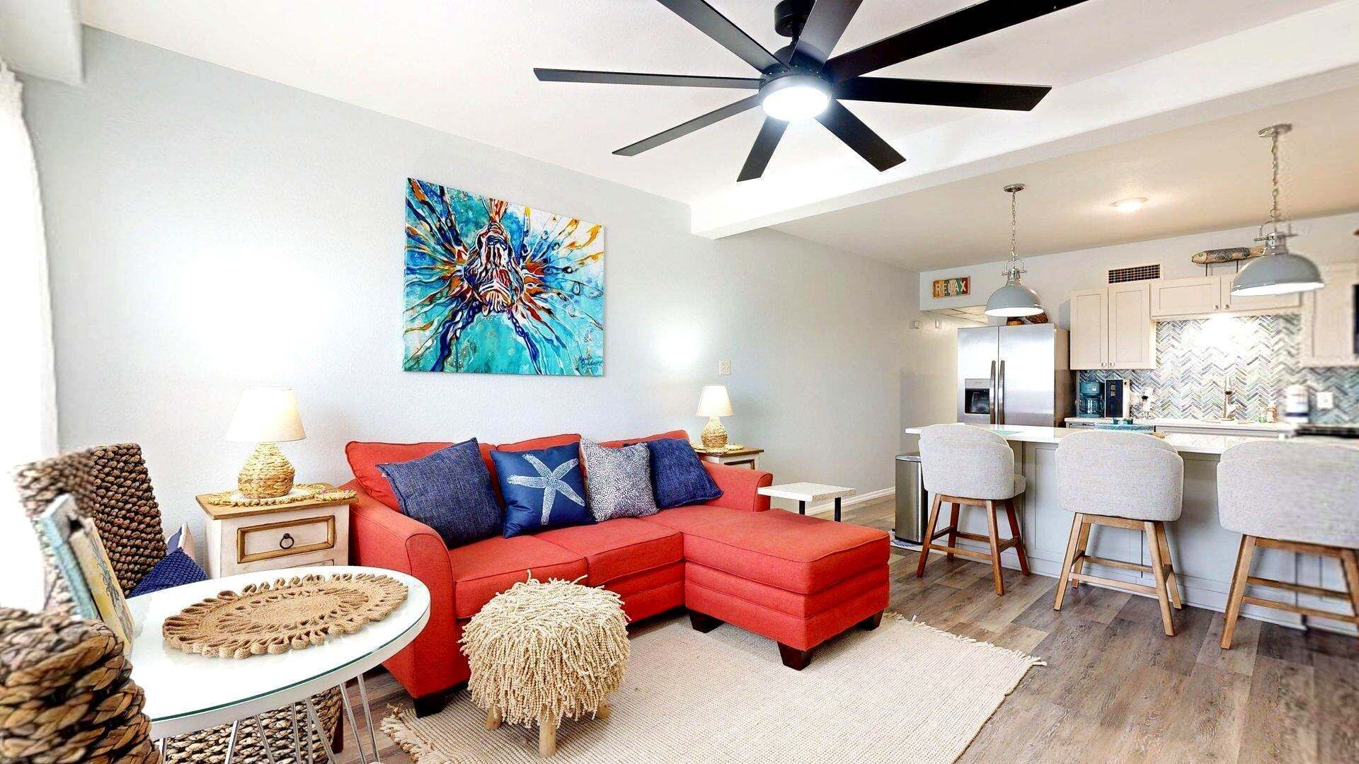 A modern living room with a red sectional sofa, a dining area with bar stools, a kitchen with a herringbone backsplash, and a ceiling fan.