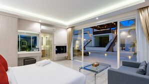Ett modernt hotellrum med en stor säng, ett soffbord i glas, en soffa, en tv och utsikt över det utomhusområde som inkluderar en trappa och en balkong.