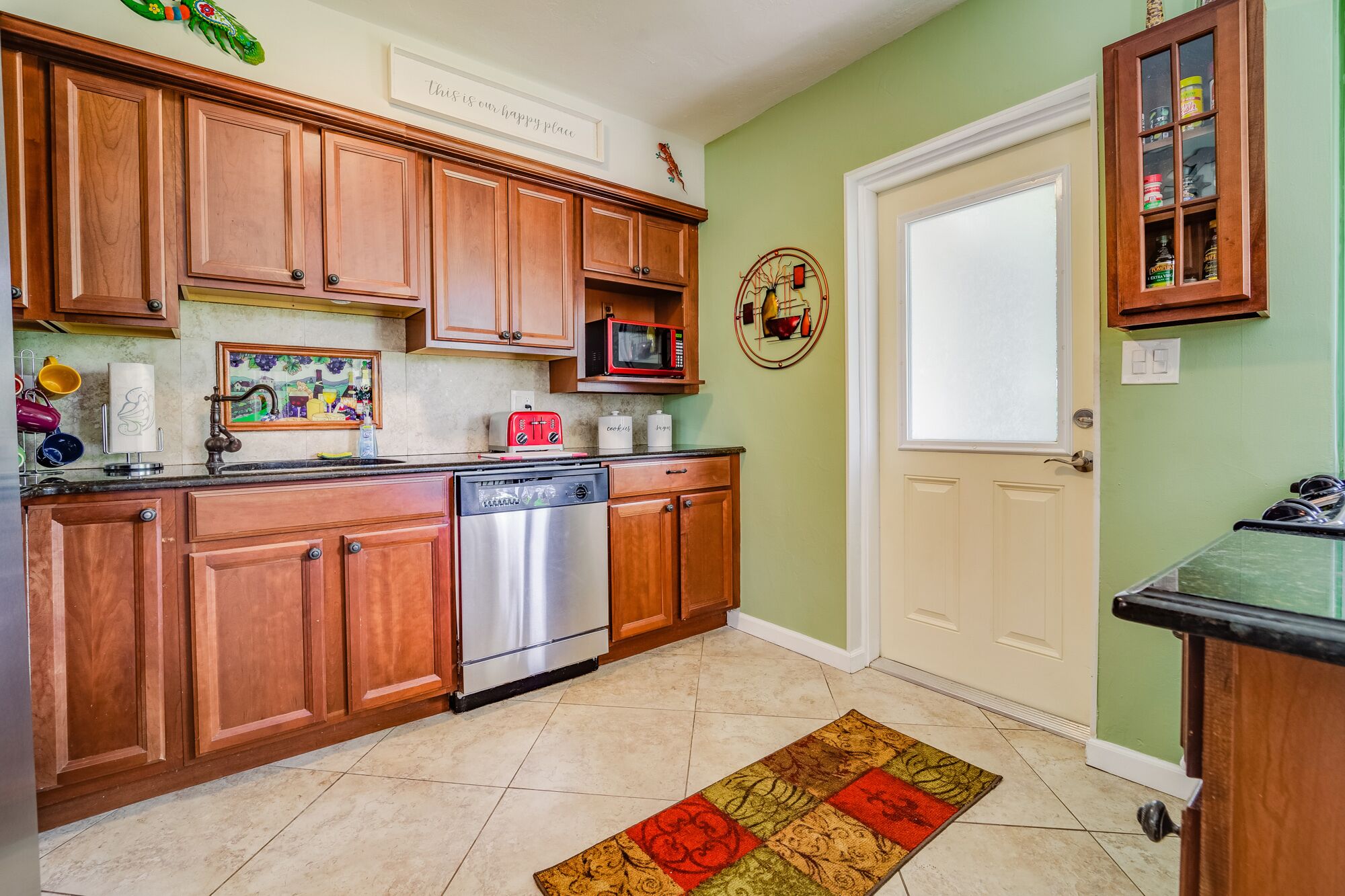 A kitchen with wooden cabinets, a stainless steel dishwasher, a microwave, and a toaster. There is a green wall and a door with a glass cabinet above it.