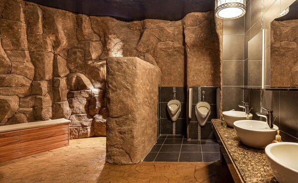 A bathroom with stone walls, two urinals, a sink, and a wooden bench. - Royal Residences by Design Union (Lagos)