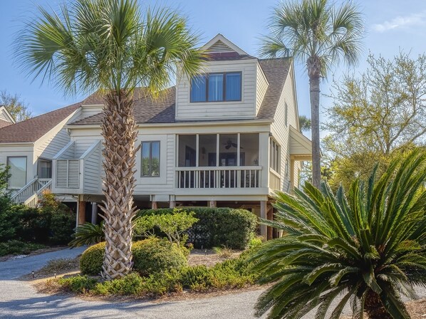A two-story house with a balcony, surrounded by palm trees and shrubs.