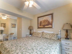 A bedroom with a large floral bedspread, a ceiling fan, a nightstand with a lamp, a chair, and a framed painting on the wall.