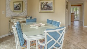 Un comedor con una mesa de vidrio rodeada de sillas blancas con cojines azules, una araña y cuadros en las paredes.