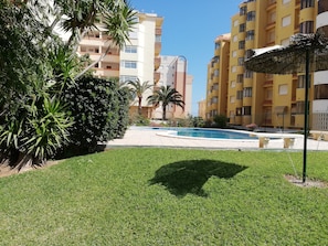 Área de piscina com céu azul, cercada por gramado bem cuidado e palmeiras, com edifícios de vários andares com varandas adjacentes.