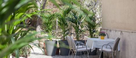 An outdoor patio with a table and chairs, surrounded by large potted plants and a concrete wall.