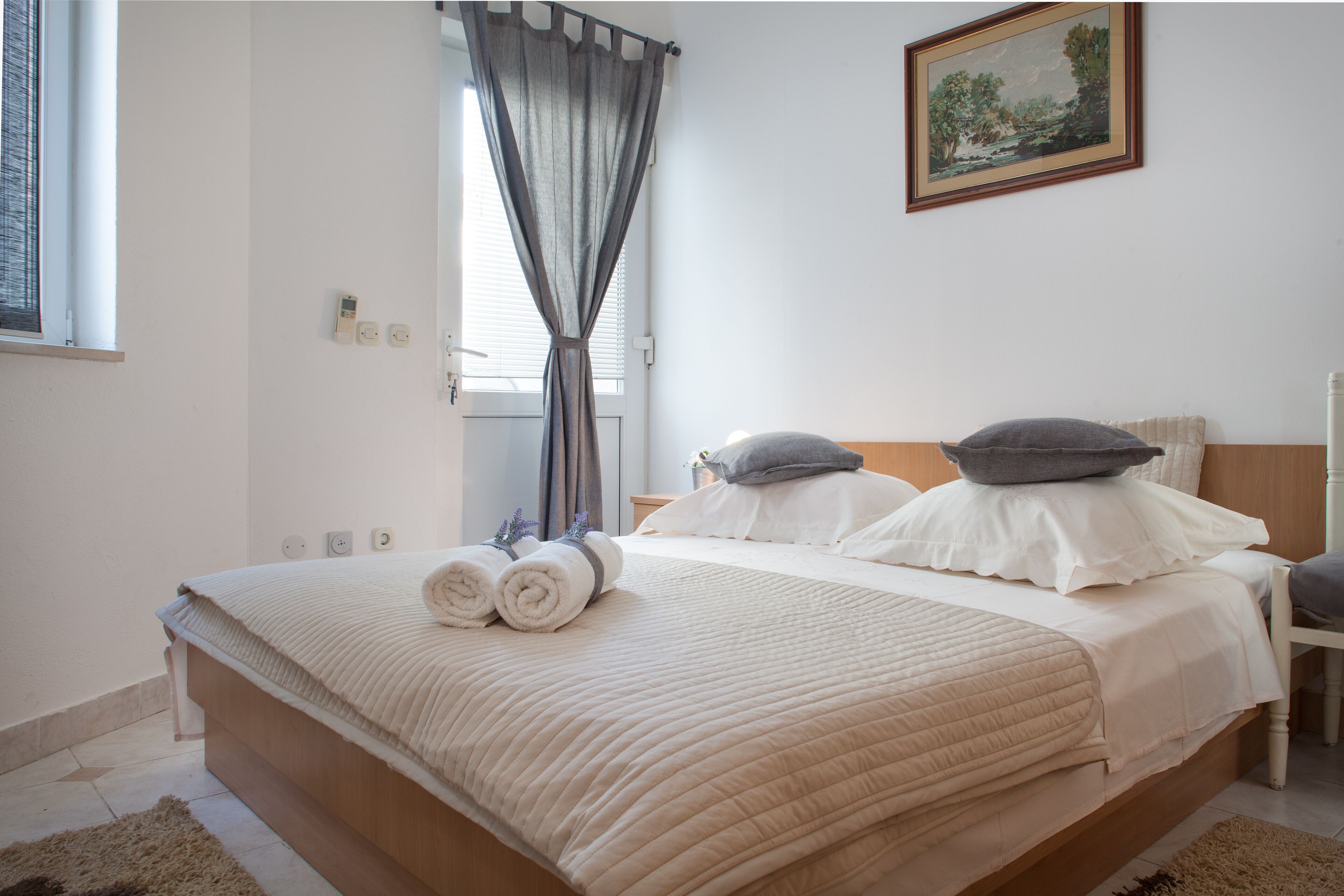 A neatly made bed with two rolled towels, a wooden headboard, and a painting on the wall.