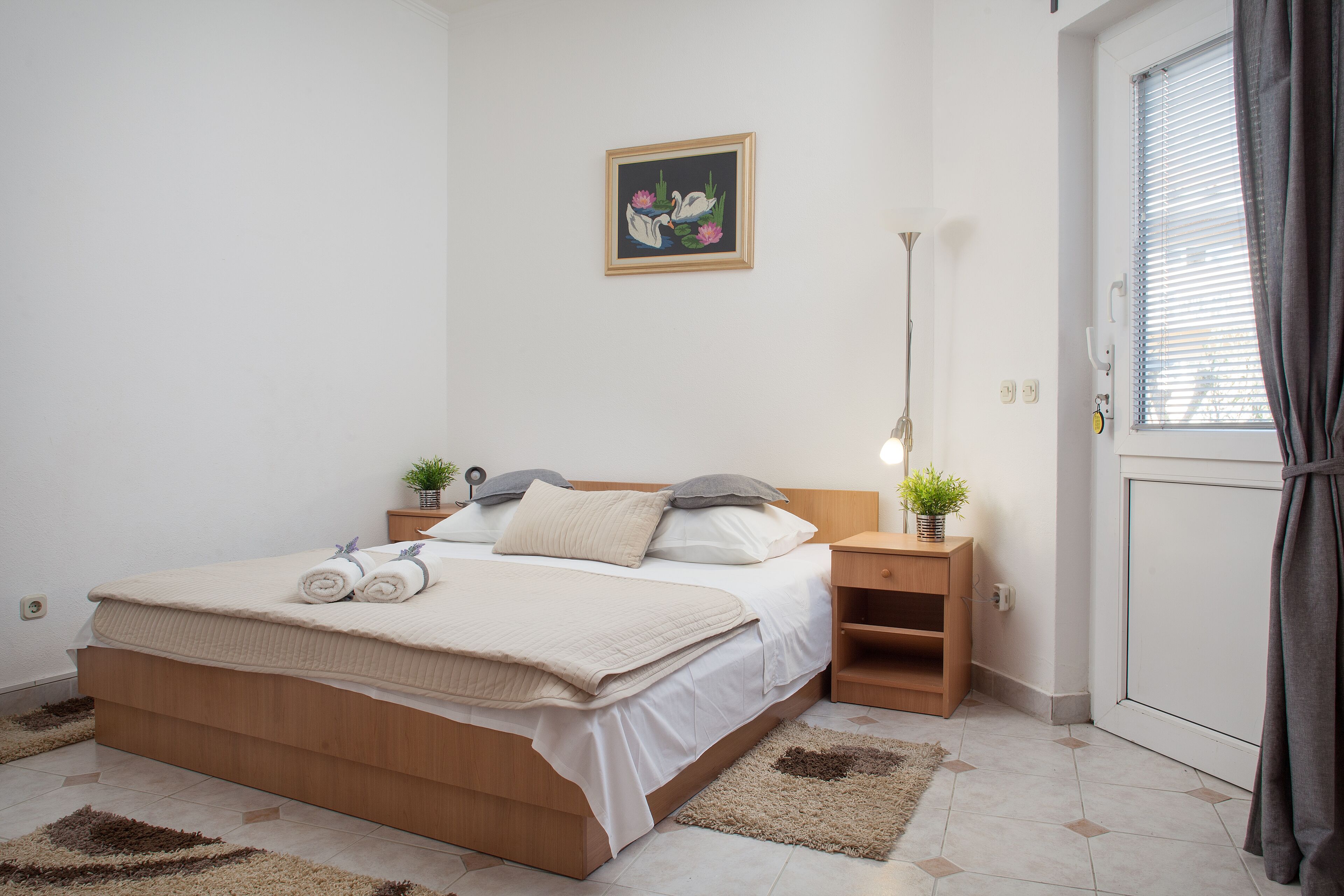 Un lit bien fait, avec des oreillers et une couverture, une table de chevet avec une lampe et une petite plante, et un tableau accroché au mur.