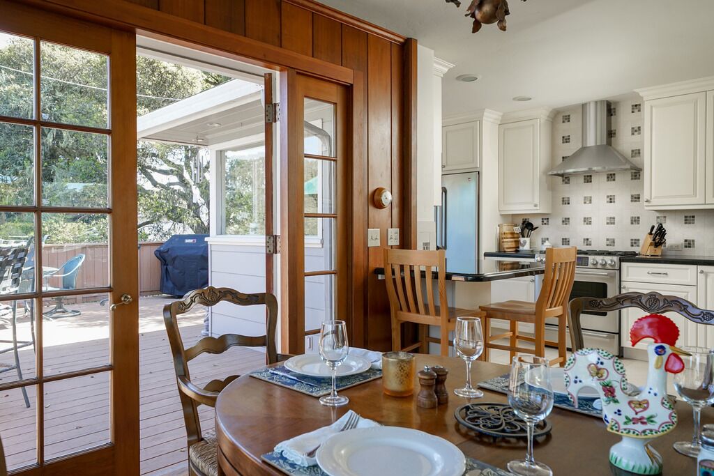 A dining area with a wooden table set for two, a kitchen in the background, and a view of the outdoors through a glass door.