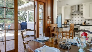 A dining area with a wooden table set for two, a kitchen in the background, and a view of the outdoors through a glass door. - Fourth Tee - Pacific Grove House - Ocean/Golf View (Pacific Grove)