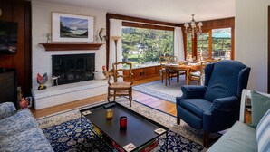 A living room with a fireplace, a dining area, and a view of greenery. - Fourth Tee - Pacific Grove House - Ocean/Golf View (Pacific Grove)