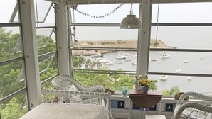 A room with a large window overlooking a body of water with boats. There is a wicker chair, a small table with a vase of flowers, and a hanging light fixture. - The View: The Name Says It All (Rockport)