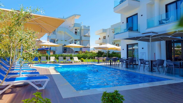 A swimming pool with lounge chairs and umbrellas, surrounded by modern buildings with balconies. - Hotel Alexander (Ksamil)