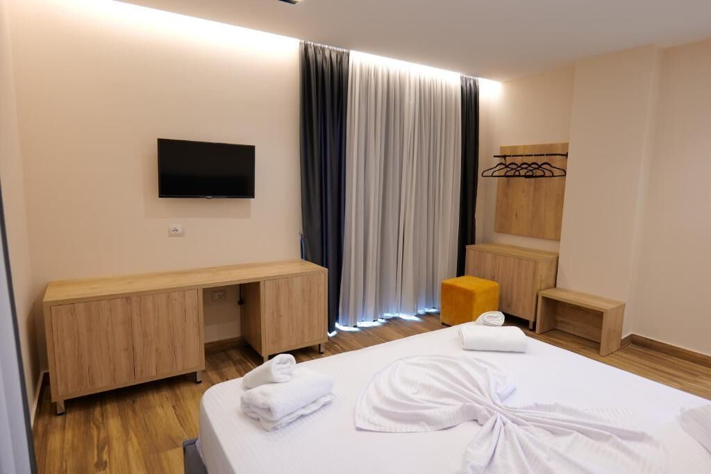 A hotel room with a bed, a TV, a wooden desk, a small wooden stool, and a wooden bench.