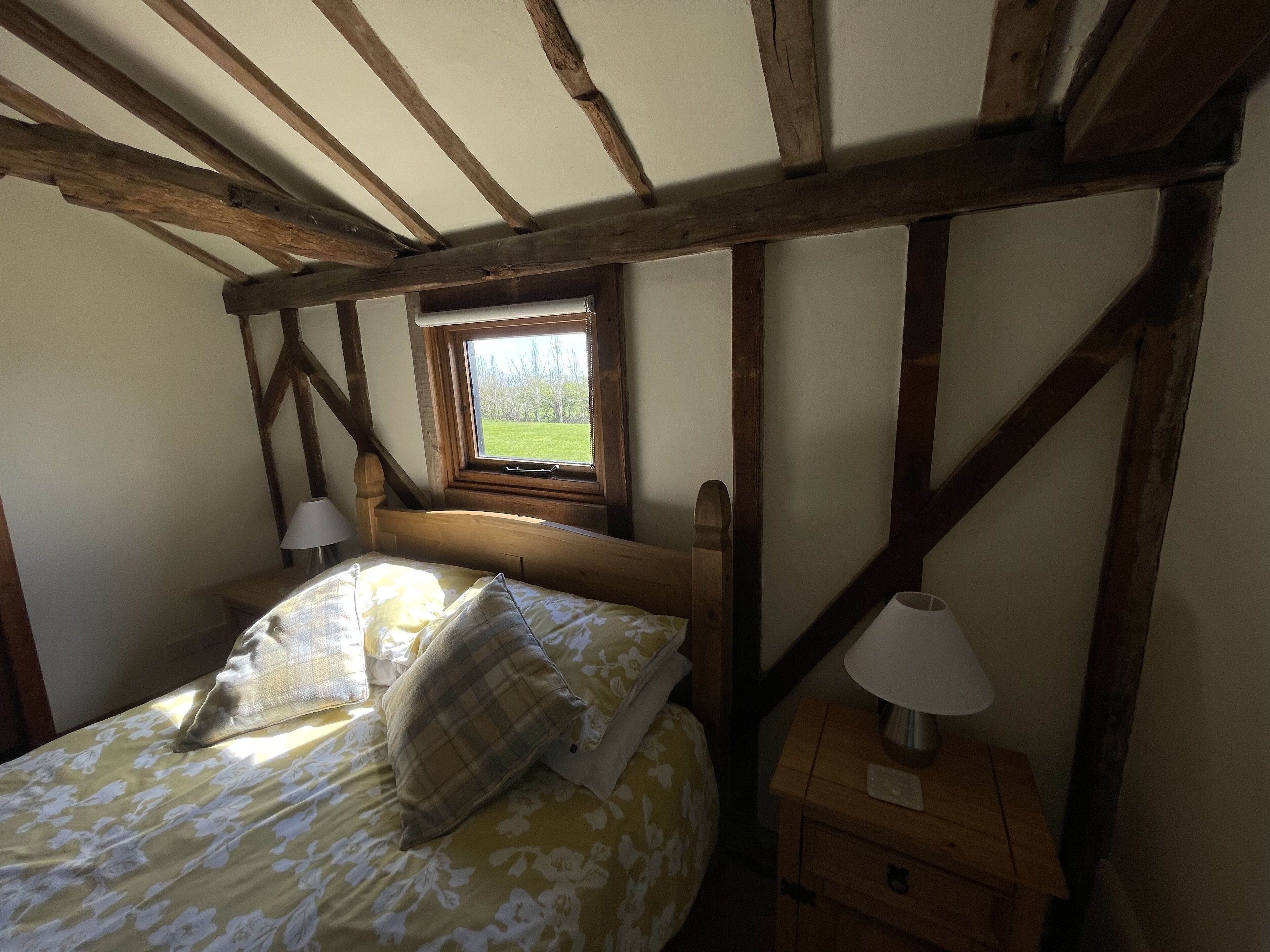Un dormitorio con techo de madera, una ventana que da a un campo verde, una cama con ropa de cama floral y dos mesitas de noche con lámparas.