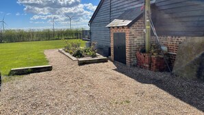 A building with a gravel driveway, a small brick structure, and a grassy area with wind turbines in the background.
