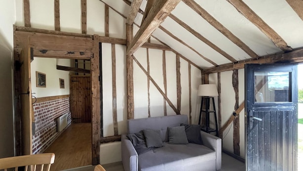 A room with exposed wooden beams, a grey sofa, a wooden chair, and a door leading to another room.