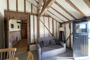 Quarto com vigas de madeira aparentes, um sofá cinzento, uma cadeira de madeira e uma porta que dá para outro cômodo.