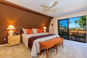 A hotel room with a large bed, two bedside tables with lamps, a wooden bench, a ceiling fan, and a balcony with a view of palm trees.