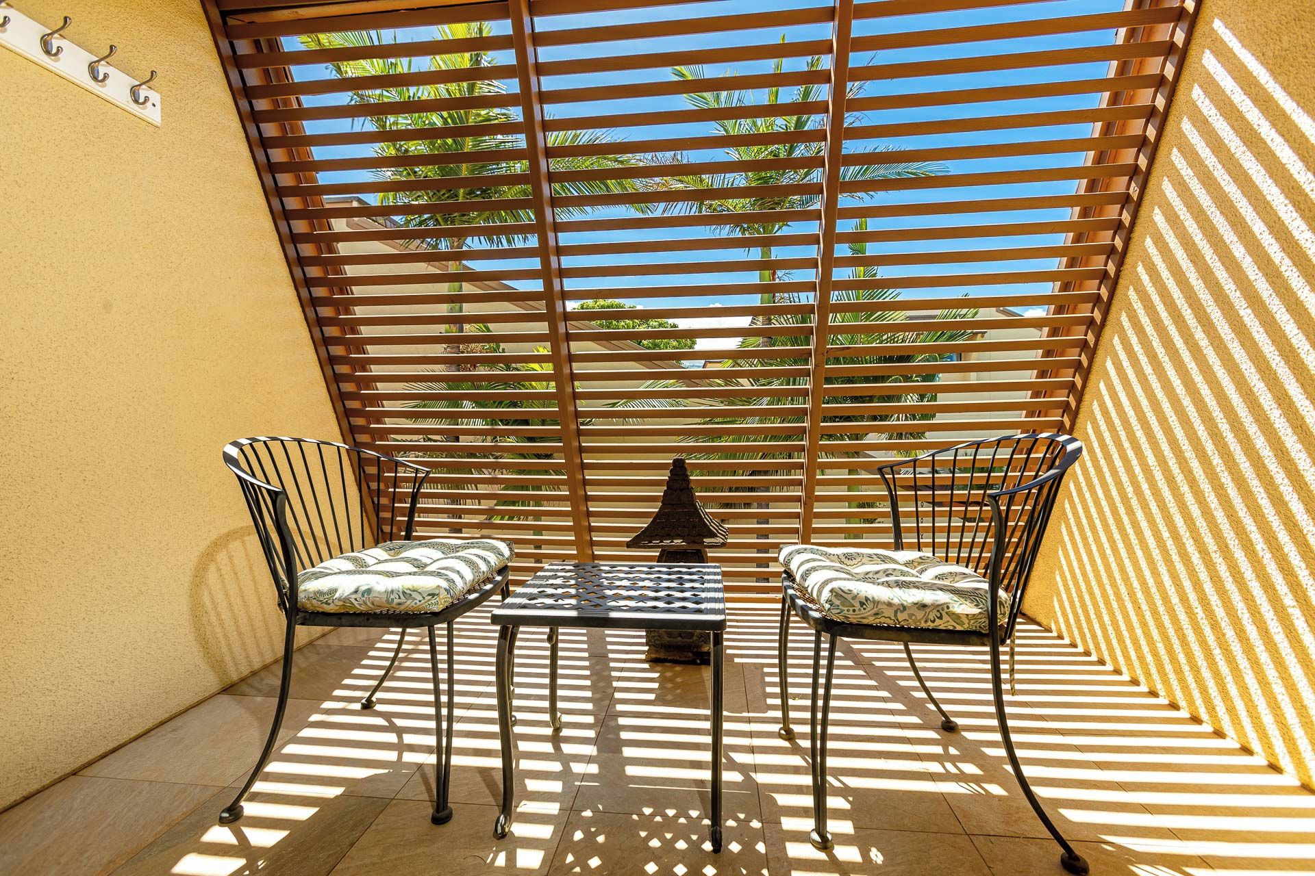 A sunlit outdoor seating area with two chairs and a table, surrounded by wooden slats providing shade.