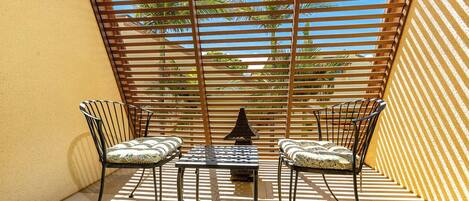 A sunlit outdoor seating area with two chairs and a table, surrounded by wooden slats providing shade.