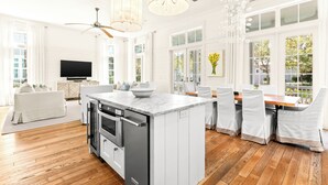 A spacious kitchen with a central island, a dining area with white chairs, a sofa, and a TV in the living area. - Chic Cottage 2 Blocks to Dragonfly Pool. With an LSV, Bikes, and More (Santa Rosa Beach)