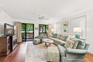 Un salon spacieux comprenant un canapé, un pouf, une table basse et un téléviseur. Il y a deux fenêtres avec des rideaux, un ventilateur de plafond et une vue sur l’extérieur.