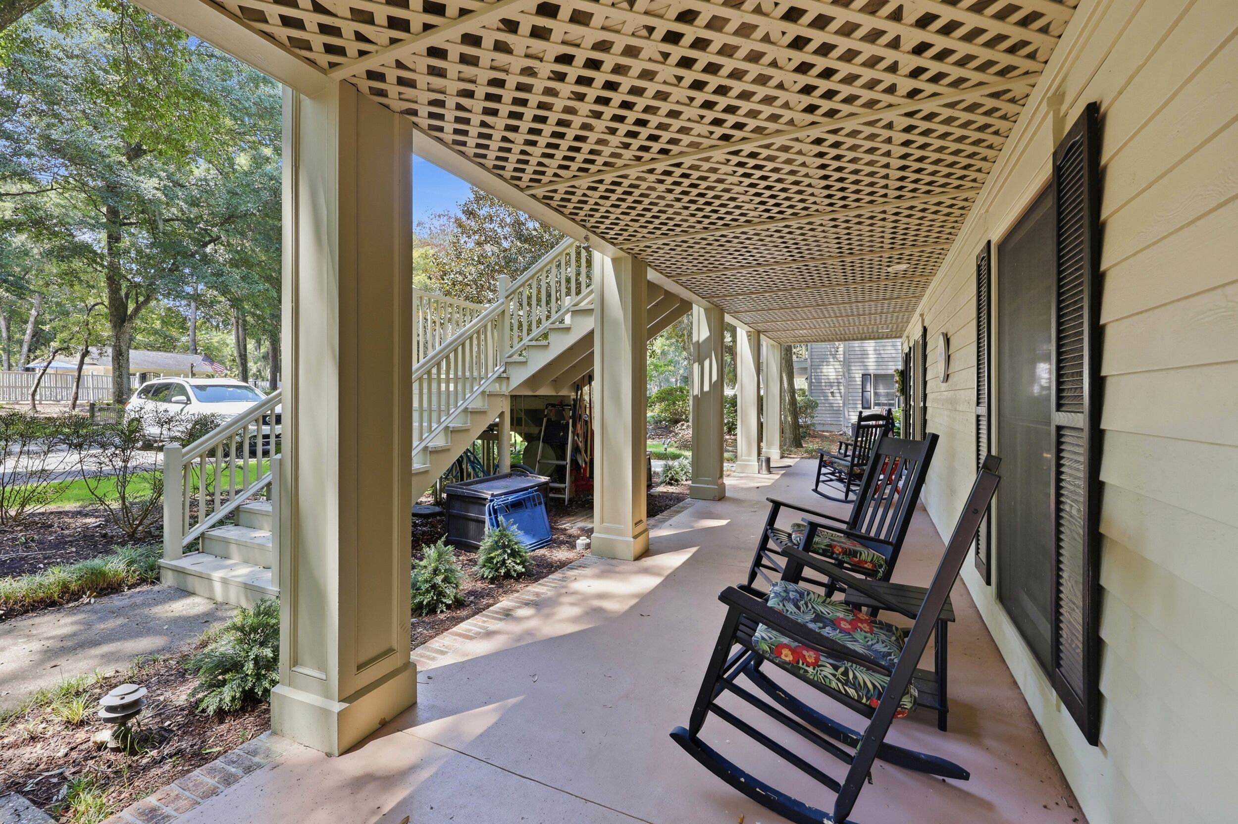 A covered porch with a staircase leading to a second level, outdoor seating, and a view of a street with parked cars.