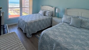 A bedroom with two beds, a ceiling fan, a nightstand with a lamp, and a view of the beach through a large window. - Find your home away from home in the Happy Landing unit (New Smyrna Beach)