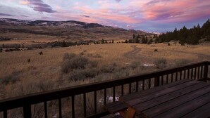 Una terraza de madera con vistas a un campo verde y montañas distantes bajo un cielo colorido.